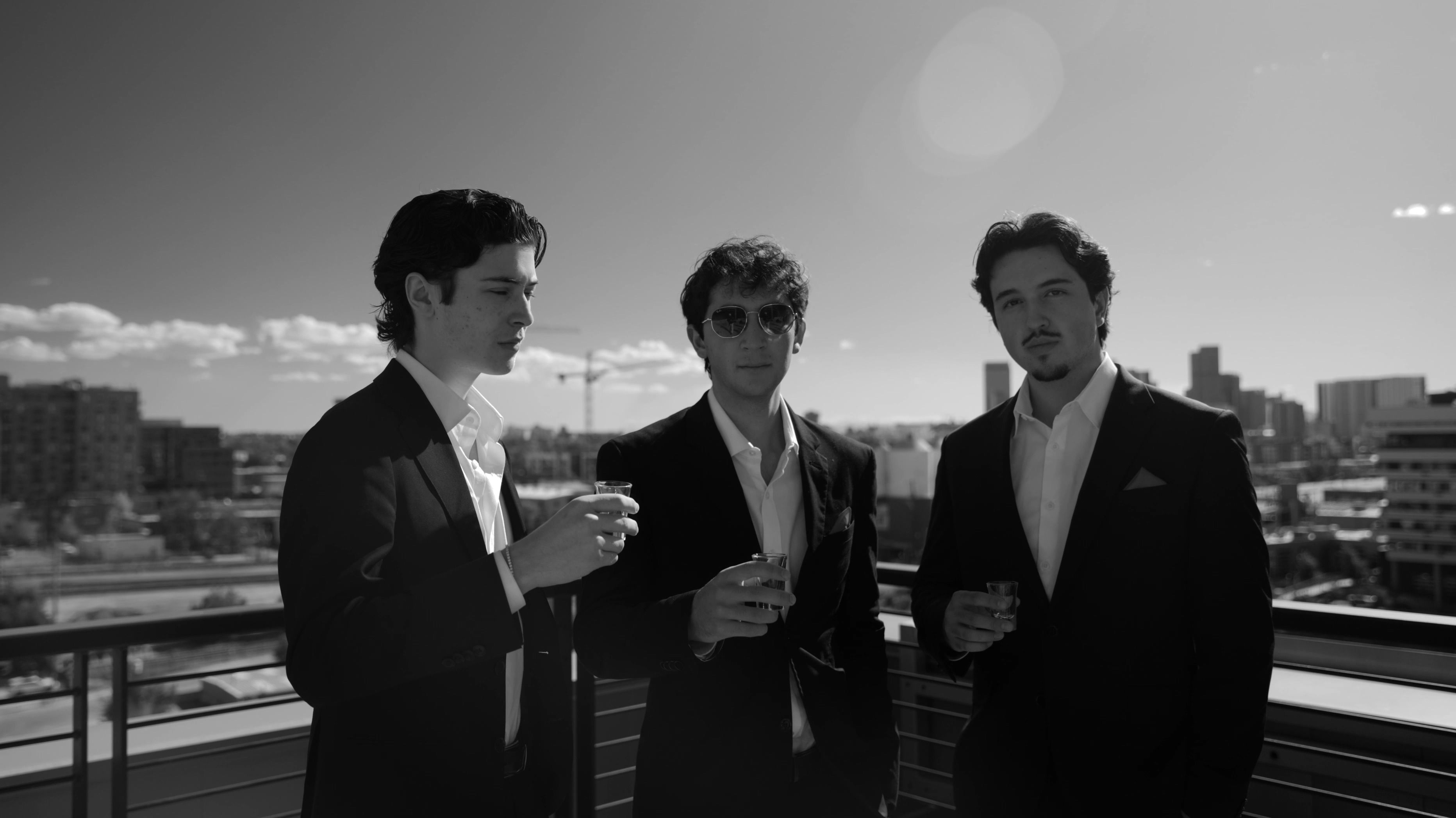 Three men in dark suits on a Denver rooftop, with the downtown skyline behind them